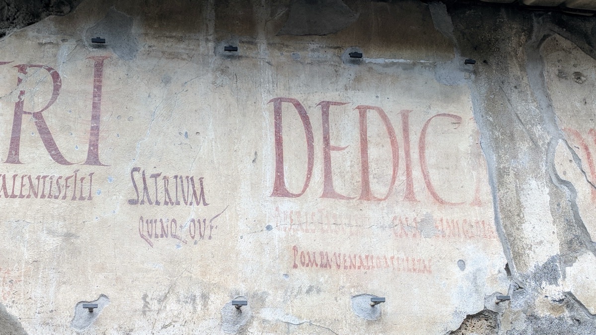 Czerwone, częściowo zatarte litery namalowane na jasnym tynku pompejańskiej ściany; widoczne fragmenty napisu i metalowe uchwyty
